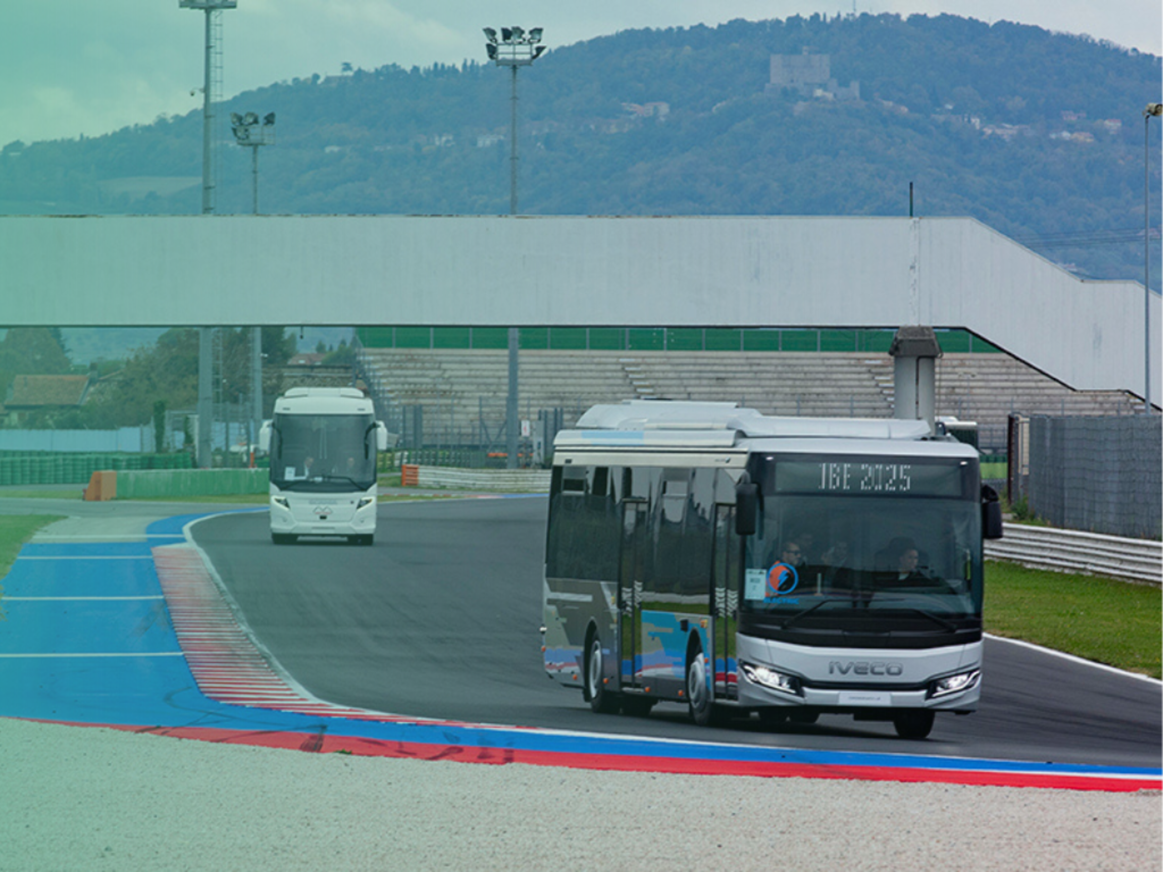 Due autobus percorrono una pista da corsa con curve blu e rosse, sullo sfondo tribune e colline con un castello; la scritta “IBE 2025” compare sul display del bus in primo piano.﻿