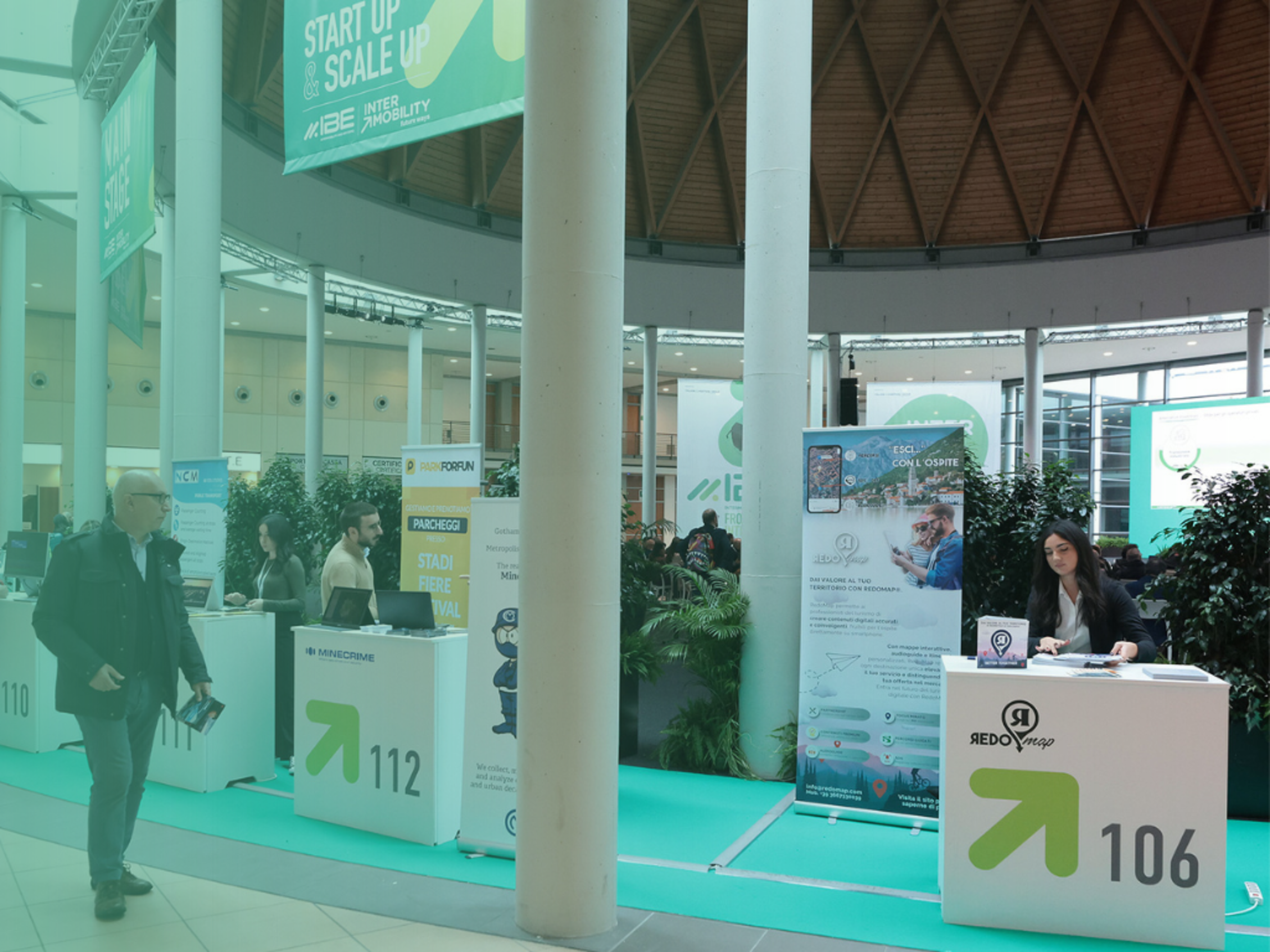 Persone agli stand informativi numerati all’interno della fiera, cartelli e banner verdi sullo sfondo.﻿