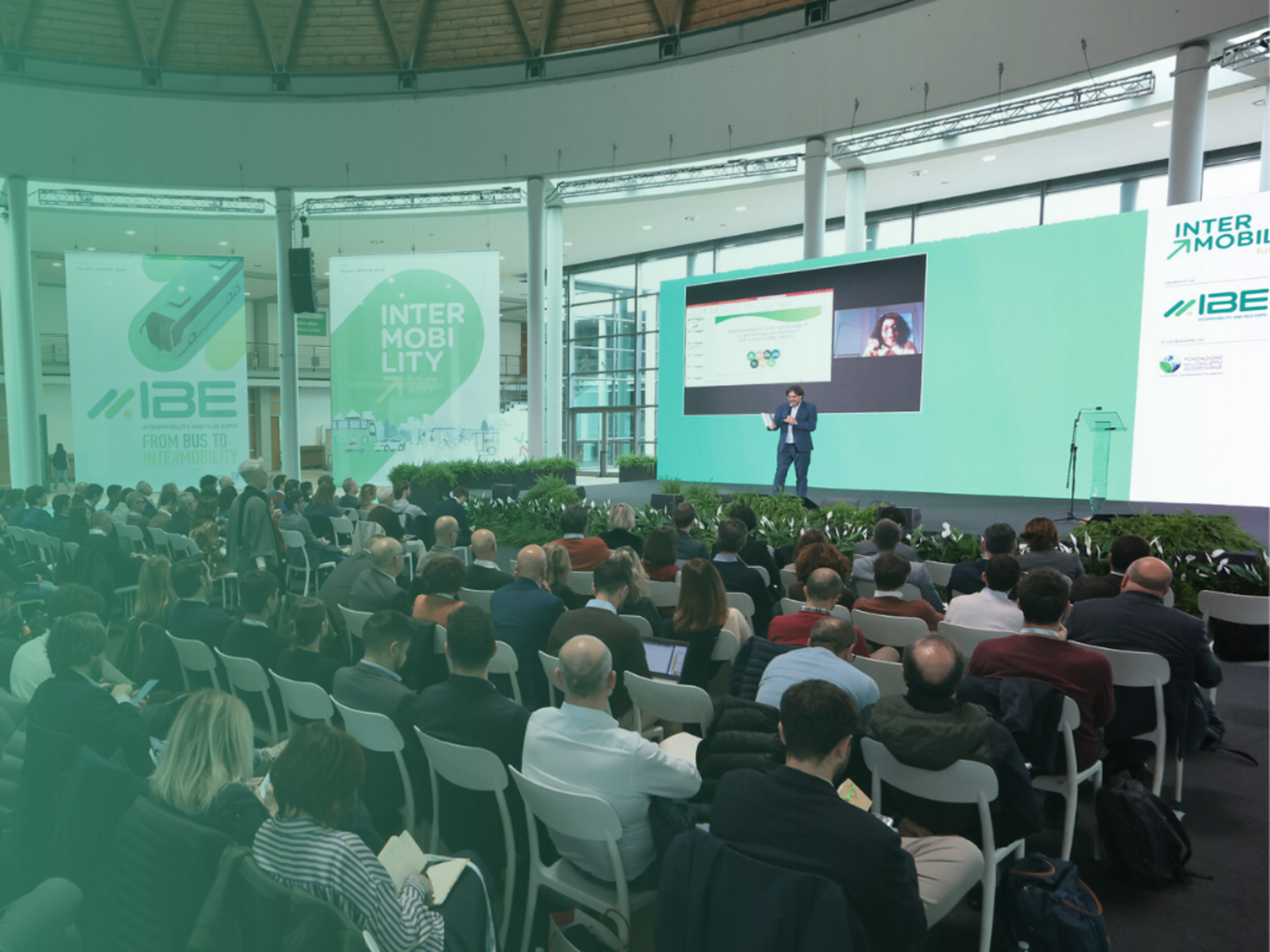Affollata platea davanti a un palco con maxischermo e relatore alla conferenza IBE, in un salone circolare con luce naturale e grandi pannelli verdi.
