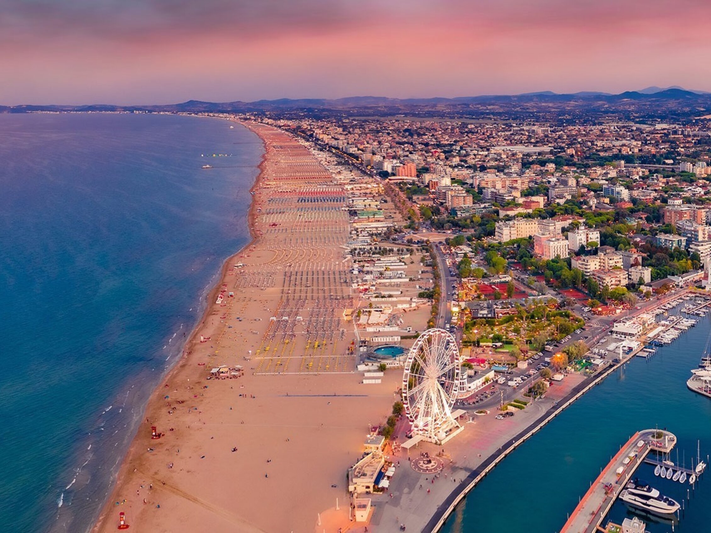 Veduta aerea della costa di Rimini al tramonto, con lunga spiaggia dorata, il porto turistico, la ruota panoramica e la città che si estende tra mare e colline sullo sfondo.