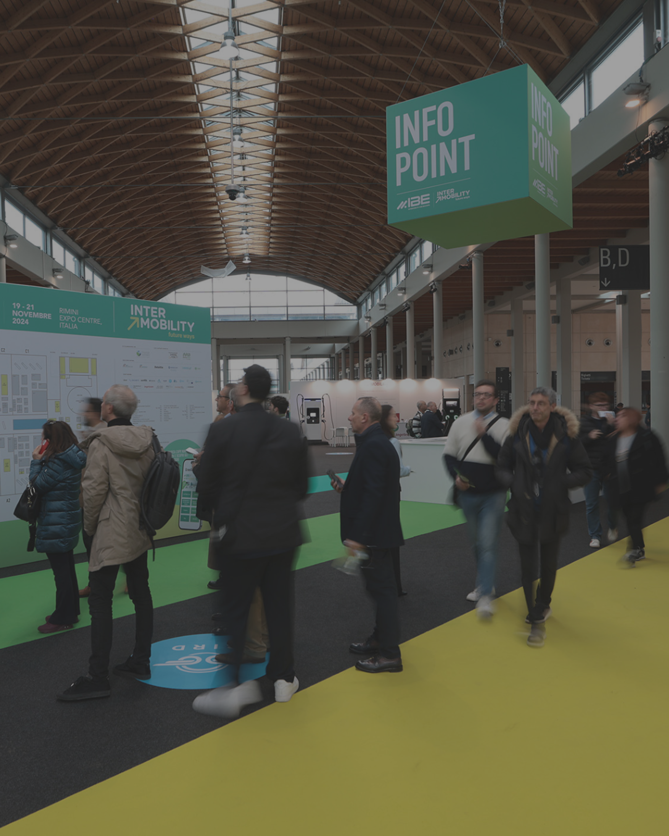 Persone in movimento nella hall di una fiera sotto un soffitto in legno; cartelli verdi 'Info Point' sospesi e mappa espositiva nell’area Intermobility.﻿