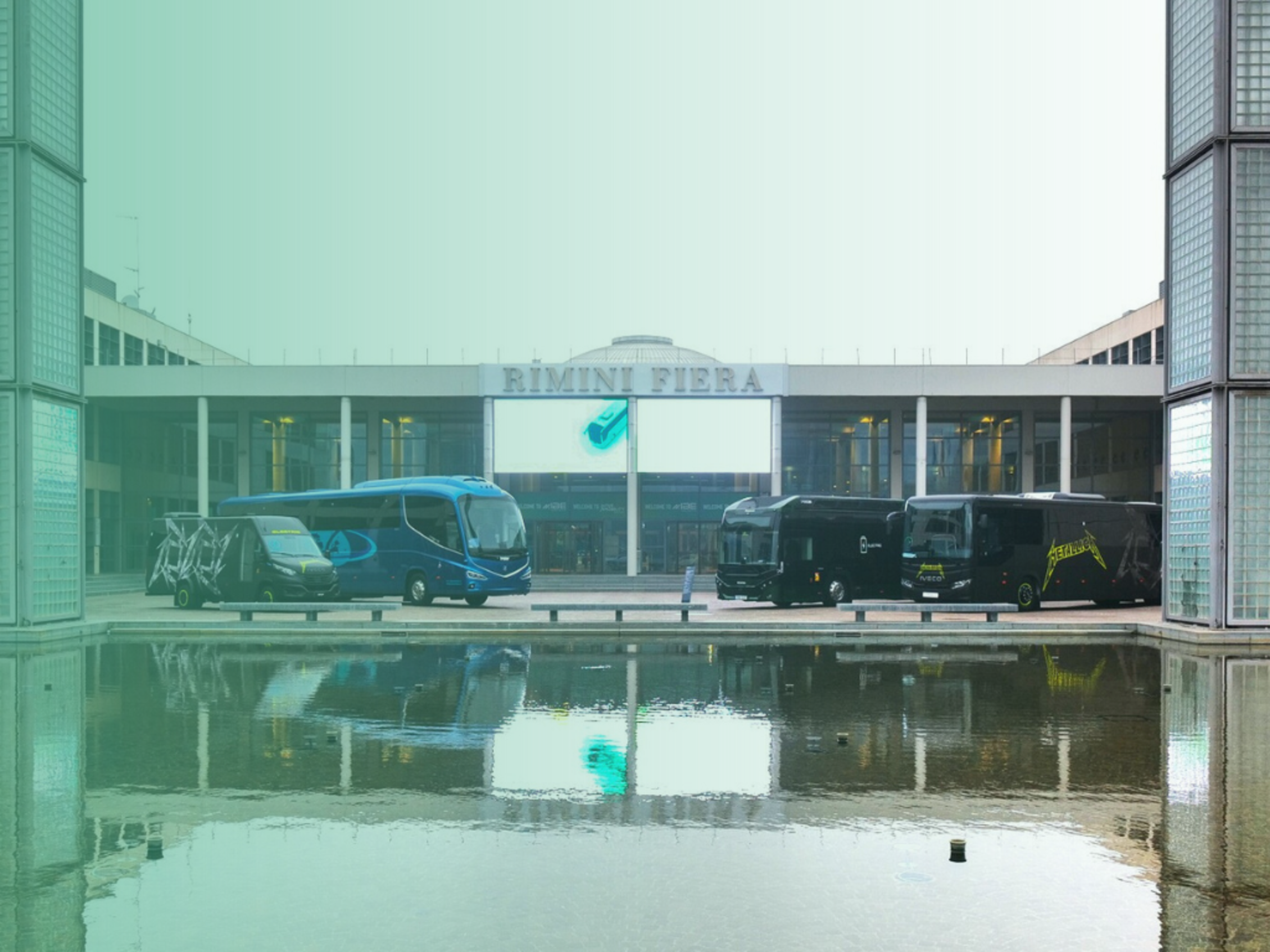 Facciata principale di Rimini Fiera con tre autobus esposti e le loro riflessioni nell’acqua antistante; l’edificio è incorniciato da colonne di vetrocemento.﻿