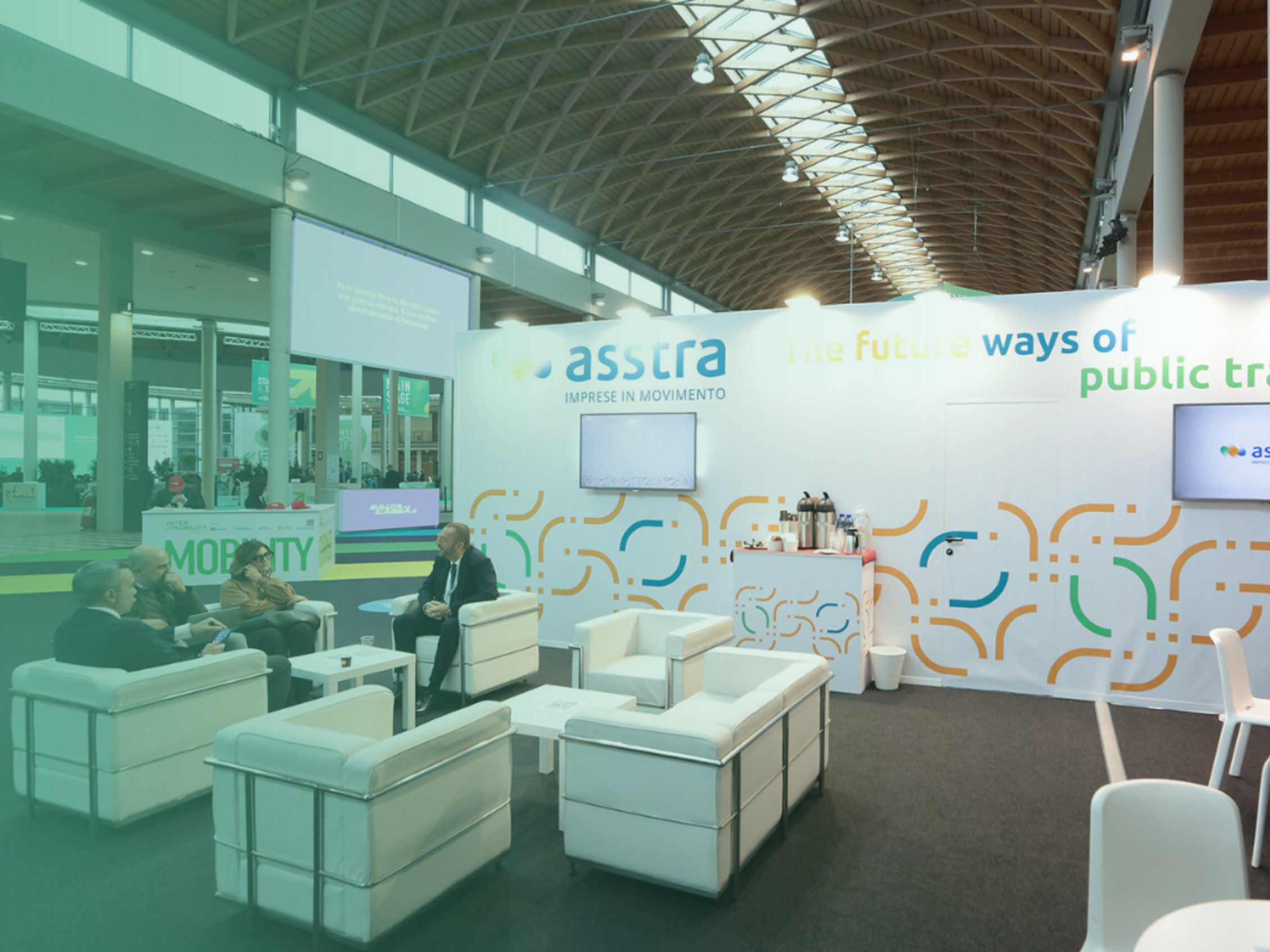 Lounge fieristico ASSTRA con quattro persone che parlano sedute su divani bianchi, parete con logo e slogan dell’associazione, ambiente moderno e ampio.﻿