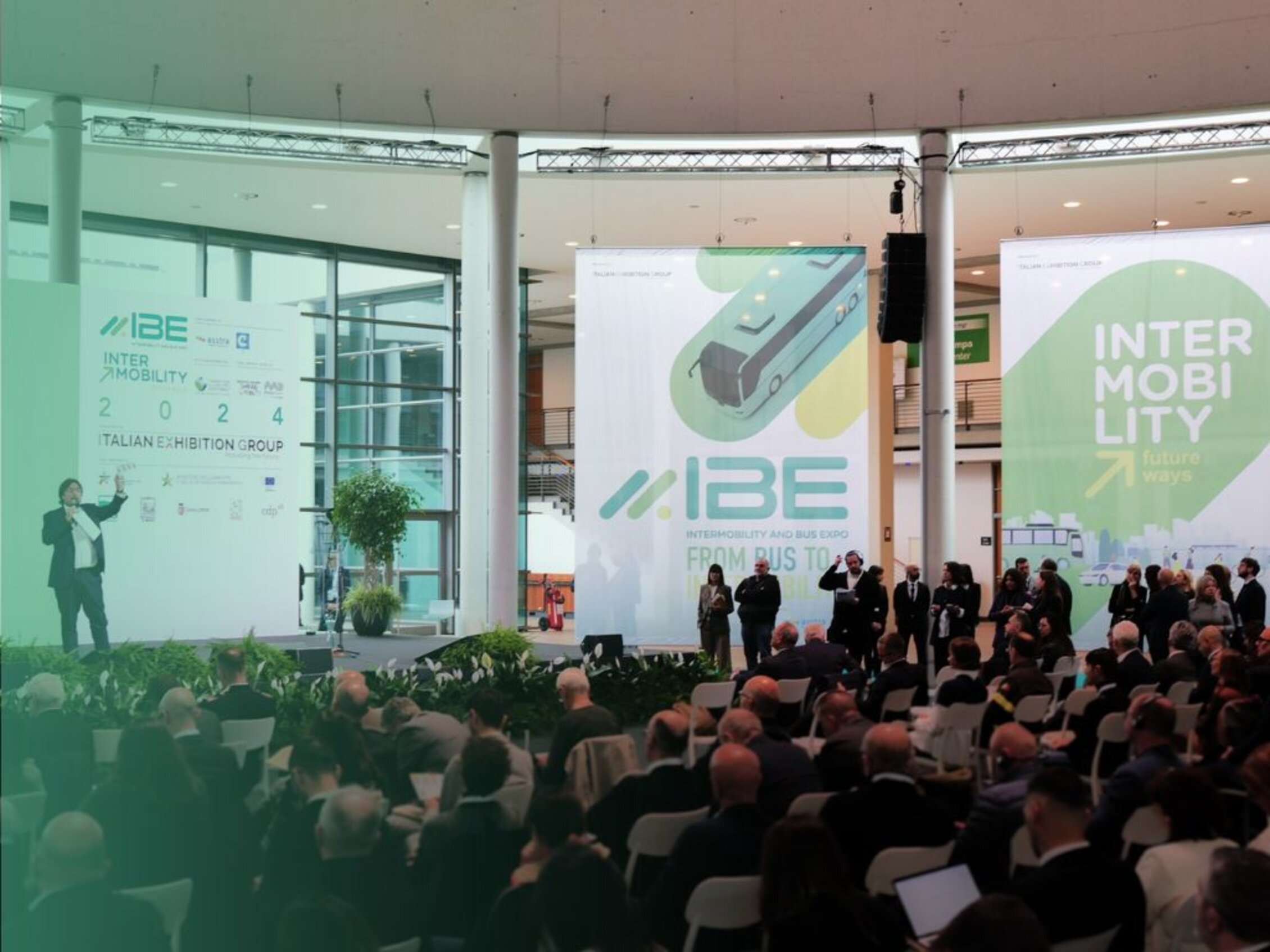 Conferenza all'interno di uno spazio fieristico moderno con pubblico seduto, relatore sul palco a sinistra, grandi poster con il logo IBE , decorazioni verdi e atmosfera professionale