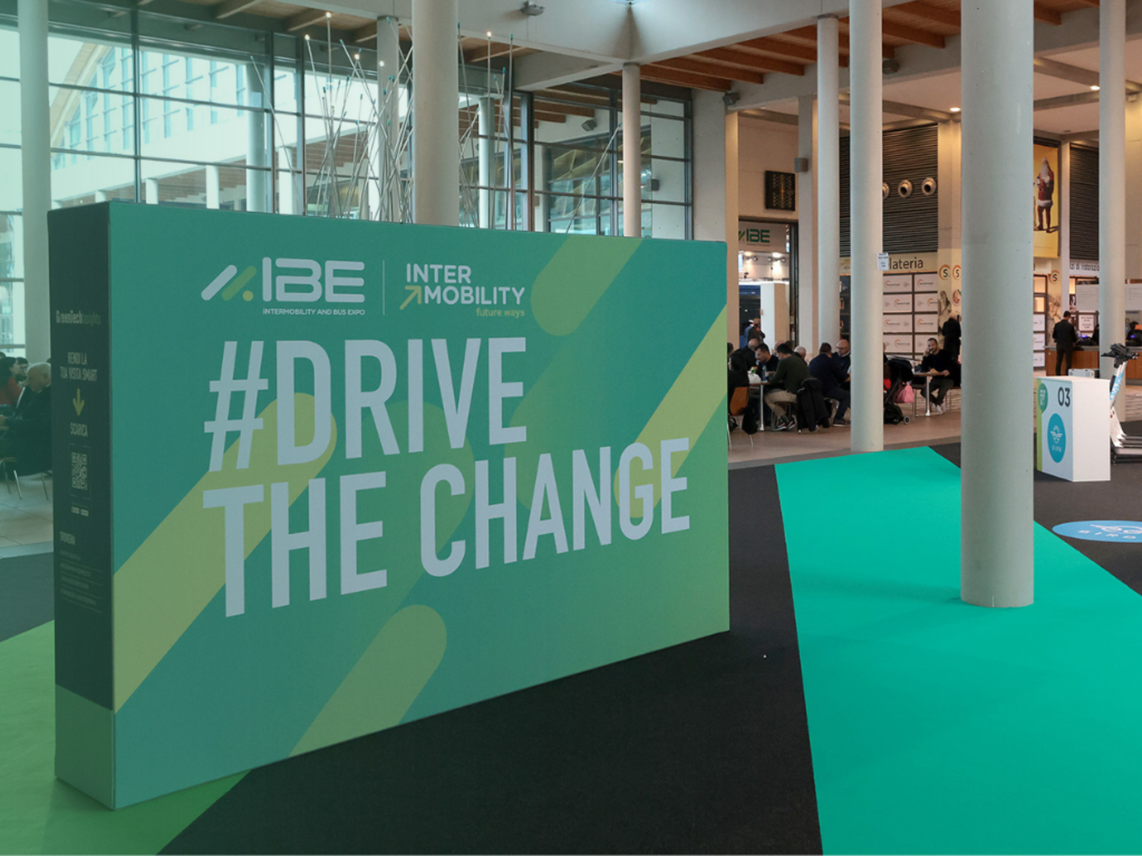 Cartellone con la scritta '#Drive the Change' alla fiera IBE Intermobility, persone sedute sullo sfondo.﻿