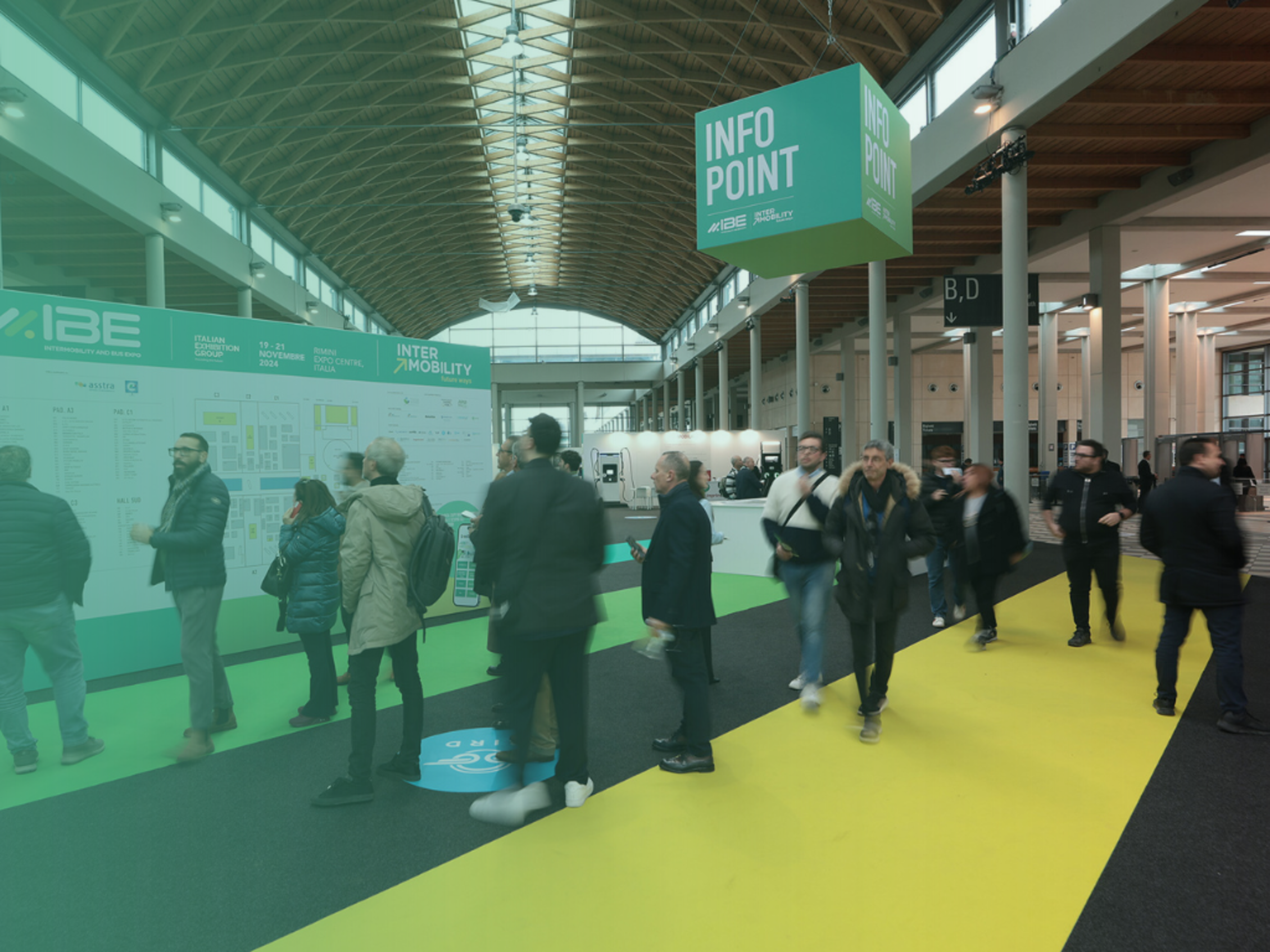 Persone camminano e osservano una mappa presso un info point in una fiera moderna.﻿